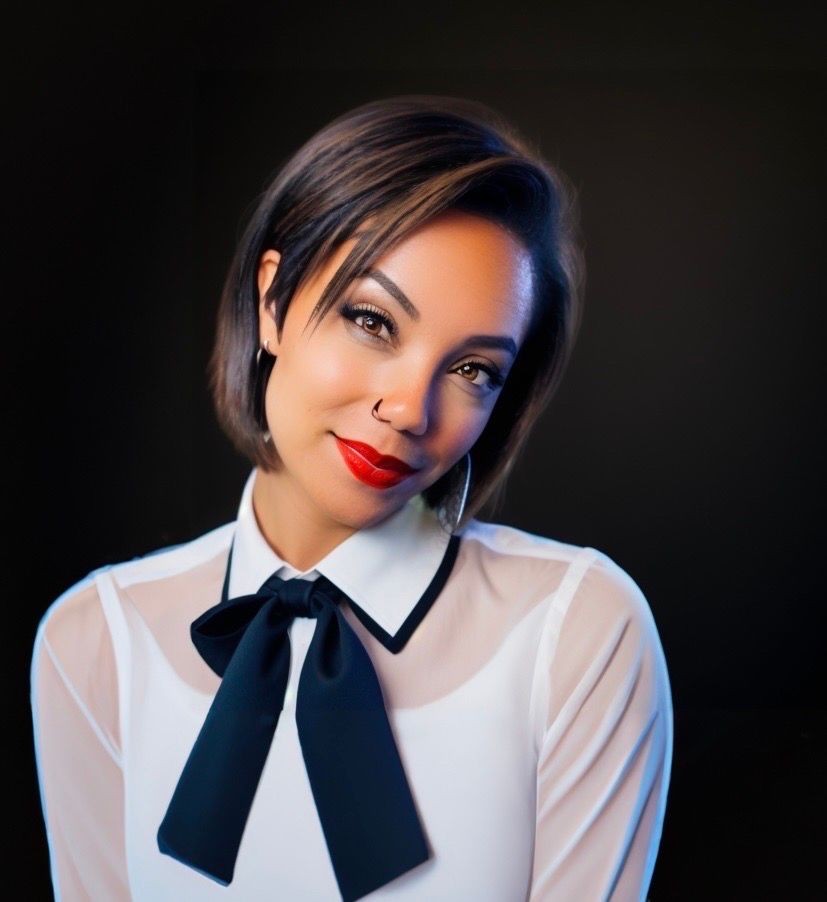 Person wearing a white blouse with sheer sleeves and a black bow tie collar against a dark background.