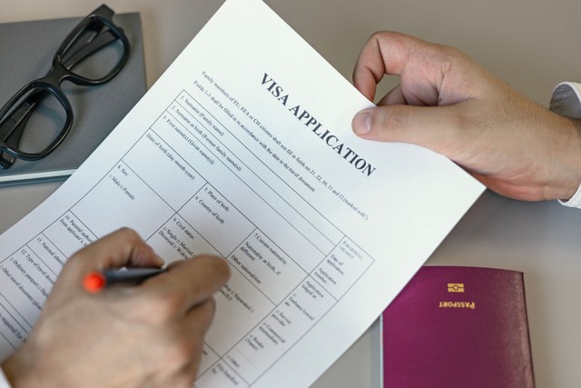 Hand holding a pen over a visa application form, with a passport on the desk
