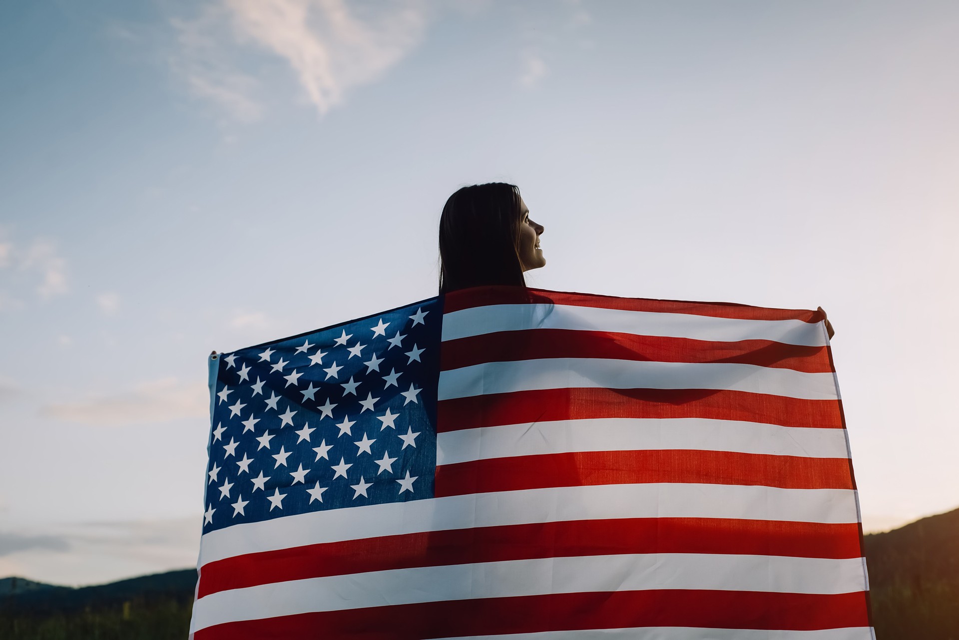 Back view close up of smile young woman waving national USA flag outdoors over sunset sky at summer in mountains, free and happy. American flag, country, patriotism, independence memorial day 4th July