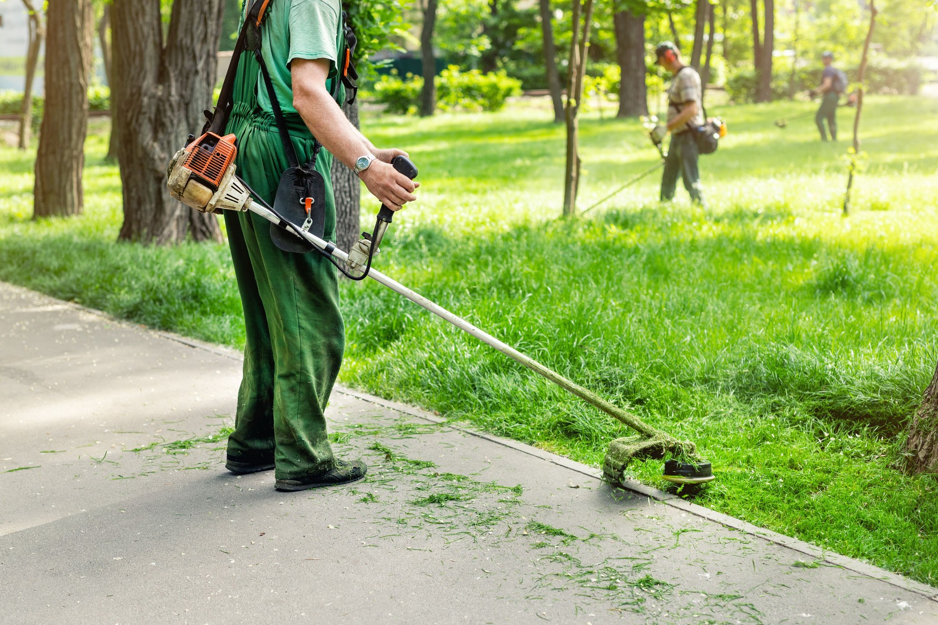 Worker mowing tall grass with electric or petrol lawn trimmer in city park or backyard. Gardening care tools and equipment. Process of lawn trimming with hand mower