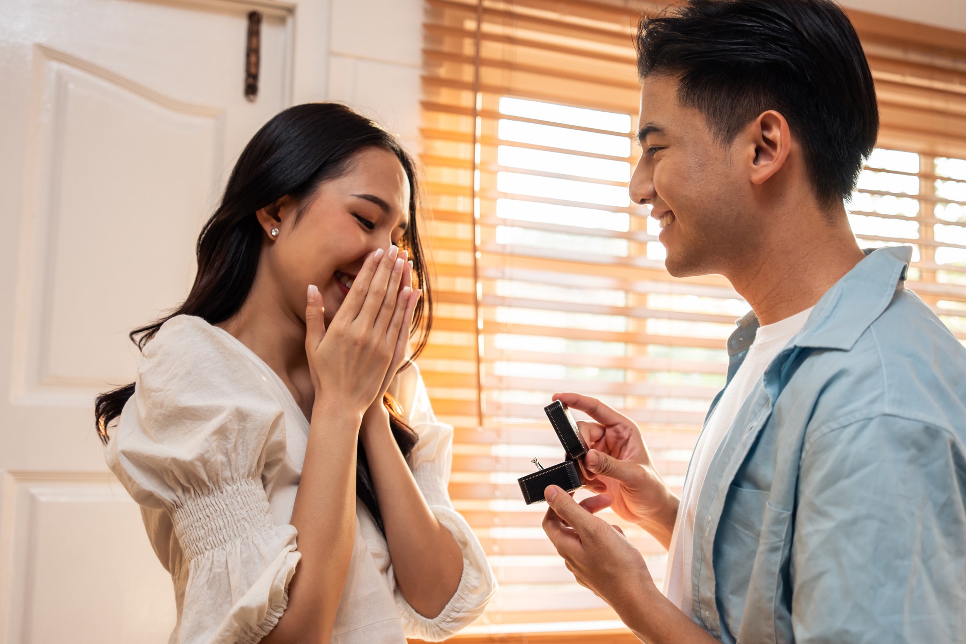 Asian romantic man making surprise proposal of marriage to girlfriend. Attractive young male and proposing to beautiful happy woman, with wedding ring enjoying surprise engagement in kitchen at home.